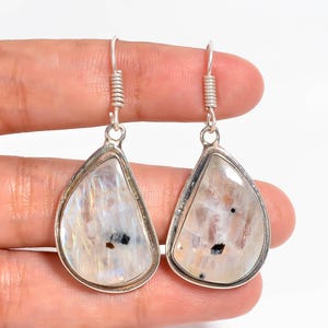 Puede incluir: Un par de pendientes en forma de lágrima con cabujones de piedra lunar blanca translúcida y pulida, engastados en metal plateado. Cada piedra muestra destellos iridiscentes internos y pequeñas inclusiones minerales oscuras y definidas.