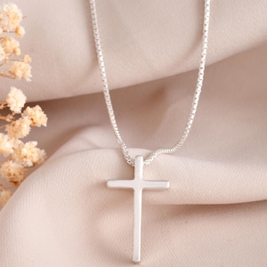 Può includere: Una collana con pendente a croce argentata. La croce è un design semplice e minimalista, di forma rettangolare. La catena è una delicata catena a scatola. Lo sfondo è di colore neutro.