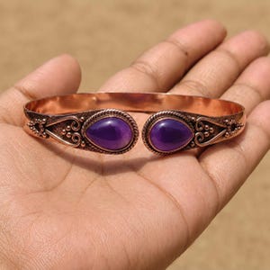 Può includere: Un bracciale rigido in rame con due pietre di ametista viola a forma di goccia alle estremità aperte. La fascia metallica presenta intricati motivi a volute in rilievo e dettagli a perline attorno a ciascuna pietra.