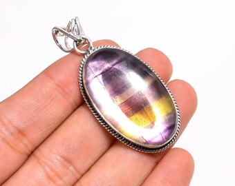 May include: Oval pendant featuring a polished fluorite gemstone with purple, yellow, and clear bands. The stone is set in a silver-colored metal frame with a textured border and a loop for a chain.