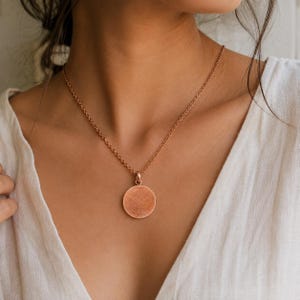 May include: A rose gold-toned necklace featuring a delicate chain and a simple, circular pendant with a textured, matte finish. The pendant hangs centrally from the chain, which rests against the collarbone area, providing a clean, metallic look.