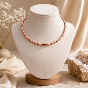Puede incluir: Un collar tipo torque circular y liso en tono oro rosa, con un diseño de banda metálica sólida y minimalista. El collar descansa sobre un busto de exhibición de color neutro, mostrando una superficie pulida y reflectante que forma un círculo completo.