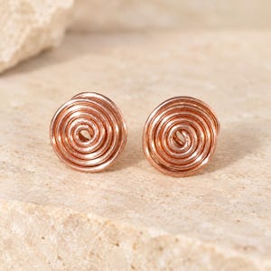 May include: A pair of rose gold-toned stud earrings featuring a circular spiral design made from coiled wire. The earrings have a polished metallic finish and a rounded, concentric pattern that radiates from the center point outward.