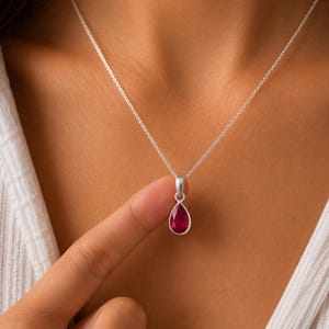 Peut inclure: Un collier en argent avec un pendentif en forme de goutte d'eau orné d'une pierre précieuse facettée d'un rouge rosé profond. Le pendentif est suspendu à une fine chaîne en argent, avec une monture en métal poli retenant la pierre translucide.