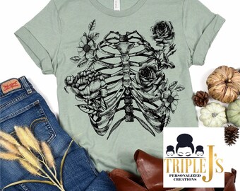 May include: A sage green t-shirt with a black and white illustration of a ribcage with flowers growing from it. The text "Triple J's Personalized Creations" is in the bottom right corner of the image.