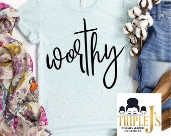 May include: A light blue t-shirt with the word "worthy" written in black cursive lettering. A cross is above the word. The shirt is on a white background with a floral scarf, a pair of jeans, and a cotton plant.