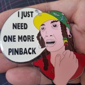 Op de afbeelding: Een witte emaille pin met een cartoonfiguur dat een gele en groene hoed en een rood shirt draagt. Het figuur houdt zijn hand aan zijn nek en zegt "Ik heb nog maar één pinback nodig".