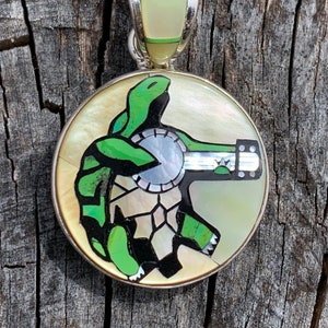 May include: A round pendant with a silver bail and a detailed design. The pendant features a green turtle playing a banjo, set against a mother-of-pearl background. The turtle and banjo are inlaid with green and black accents.