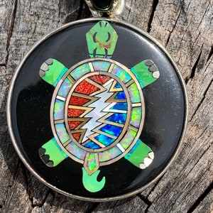 May include: A round pendant with a black background and a turtle design. The turtle has green limbs and head, with a colorful shell featuring a lightning bolt and other geometric shapes. The pendant has a silver border and bail.
