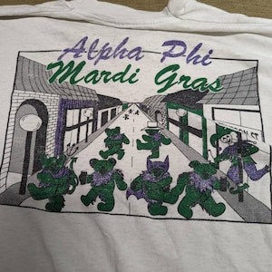 May include: White t-shirt with a graphic print of a street scene with the text "Alpha Phi Mardi Gras" and cartoon bears in costumes.