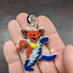 May include: A colorful pendant in the shape of a dancing bear. The bear has a red head, a blue body, and a white lightning bolt design. The pendant is made of inlaid materials and has a silver-colored bail.