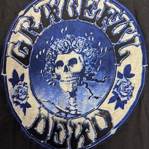 May include: A blue and white circular graphic with the text "Grateful Dead" in a black, stylized font. The graphic features a skull with a wreath of roses and a skeletal figure. The text "1976 Kelly-Mouse TM G.D.A." is printed below the graphic.
