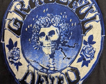 May include: A blue and white circular graphic with the text "Grateful Dead" in a black, stylized font. The graphic features a skull with a wreath of roses and a skeletal figure. The text "1976 Kelly-Mouse TM G.D.A." is printed below the graphic.