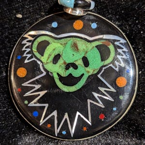 May include: A round pendant with a black background featuring a green bear design. The bear has a smiling face and is surrounded by a silver zig-zag border. The pendant has a silver bail with turquoise and black accents.