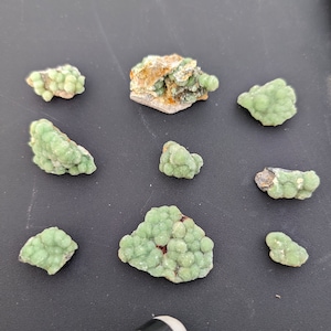 May include: A collection of nine light green mineral specimens with a bubbly, clustered texture. Some pieces have a brown matrix. The minerals are arranged on a dark gray surface. The image is well-lit, highlighting the color and texture of the stones.