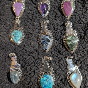 May include: Nine silver cat pendants with various gemstone hearts. The gemstones include amethyst, turquoise, moonstone, and green aventurine.
