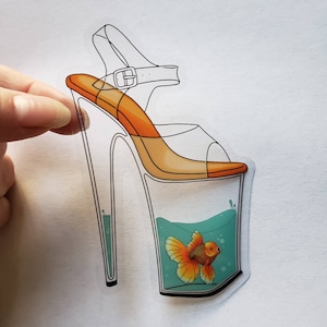 May include: A clear high-heeled shoe with an orange platform and a goldfish swimming inside. The shoe is outlined in black and the goldfish is orange and white.