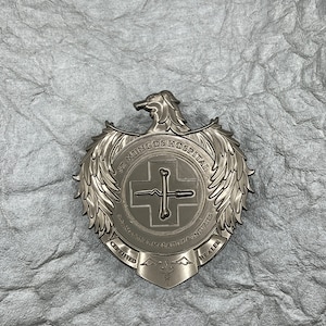 Peut inclure: Un badge en métal argenté avec un design d'aigle ailé. Le badge porte l'inscription "St. Mungo's Hospital" et "Healer of Maladies & Injuries". Il indique également "Certified" et "Healer".