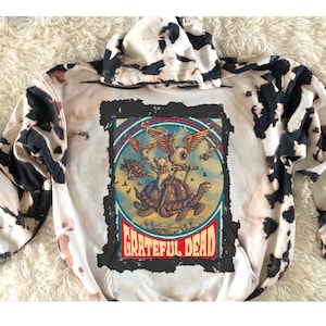 May include: A white and black tie-dye hoodie with a vintage Grateful Dead graphic featuring a cat riding a turtle with wings. The graphic is in a distressed, vintage style with red, blue, and yellow colors.
