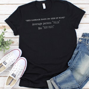 May include: Black t-shirt with white text that reads "*SEES GARBAGE BAGS ON SIDE OF ROAD* Average person: "TRASH" Me: "BODY PARTS"