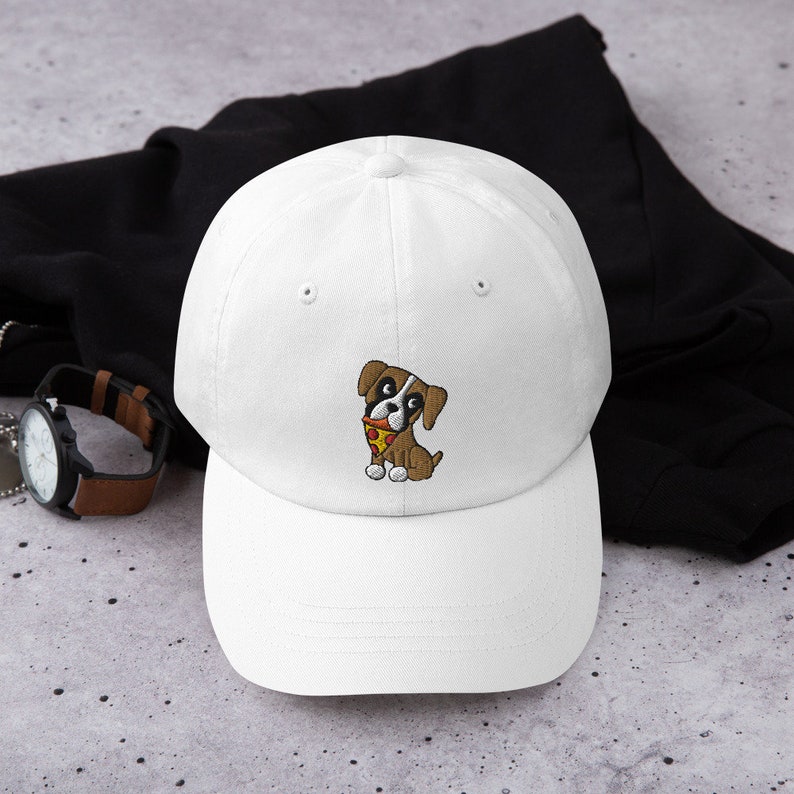 May include: White baseball cap with a curved brim. The cap features an embroidered cartoon dog with brown and white fur, holding a slice of pizza. The dog is wearing a red collar. The cap is a classic, casual accessory.