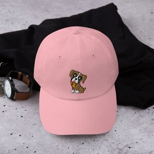 May include: A light pink baseball cap featuring an embroidered cartoon dog holding a slice of pizza. The dog is brown and white with black eyes. The cap is made of a soft material and has a curved brim.