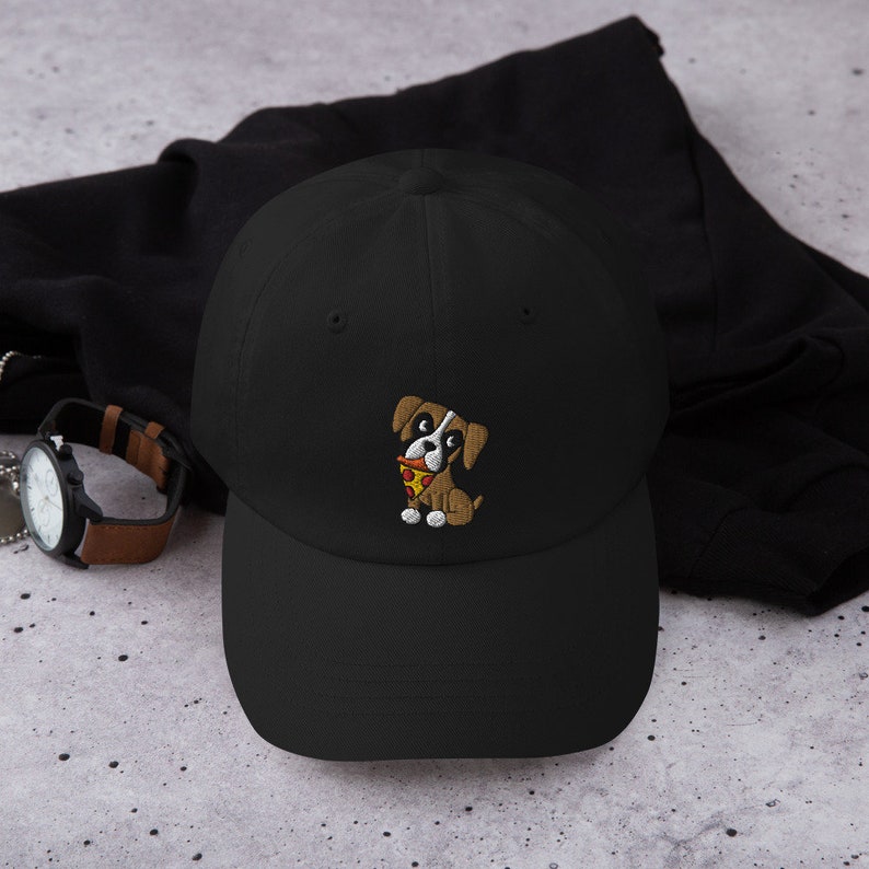 May include: Black baseball cap with an embroidered cartoon dog holding a slice of pizza. The dog is brown and white with a red collar. The cap is made of a soft material and has a curved brim.