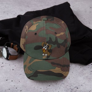 May include: Camouflage baseball cap with an embroidered cartoon dog holding a slice of pizza. The cap features a brown, green, and tan pattern. A watch with a brown leather band is visible in the background.