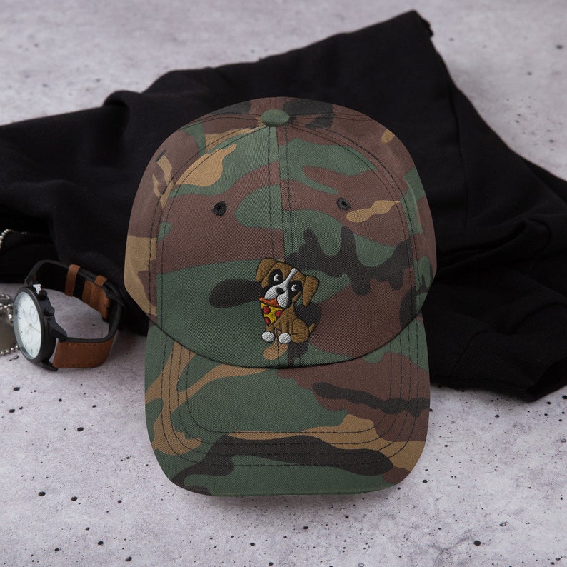 May include: Camouflage baseball cap with an embroidered cartoon dog holding a slice of pizza. The cap features a brown, green, and tan pattern. A watch with a brown leather band is visible in the background.