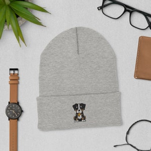 Puede incluir: Gorro gris con un perro negro y blanco bordado en la parte delantera. El perro lleva un hueso amarillo en la boca.