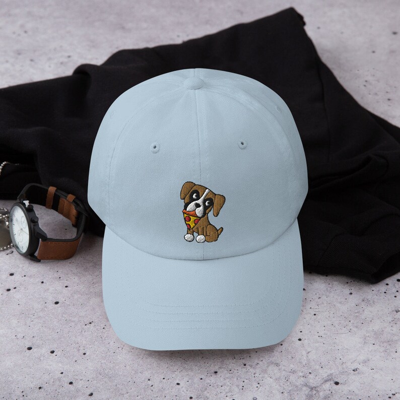 May include: A light blue baseball cap featuring an embroidered cartoon dog holding a slice of pizza. The dog is brown and white with black eyes. The cap has a curved brim and a six-panel design.