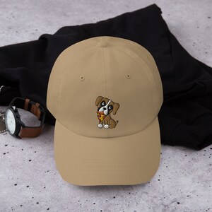 May include: A tan baseball cap featuring an embroidered cartoon dog with a slice of pizza. The dog is brown, white, and black, with a red collar. The cap is made of a soft material and has a curved brim.