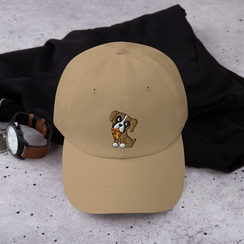 May include: A tan baseball cap featuring an embroidered cartoon dog with a slice of pizza. The dog is brown, white, and black, with a red collar. The cap is made of a soft material and has a curved brim.