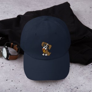 May include: A navy blue baseball cap featuring an embroidered cartoon dog holding a slice of pizza. The dog is brown and white with a red collar. The cap is made of a soft material and has a curved brim.