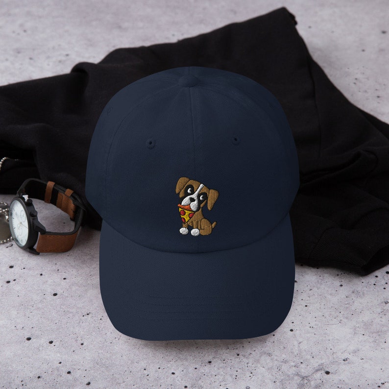 May include: A navy blue baseball cap featuring an embroidered cartoon dog holding a slice of pizza. The dog is brown and white with a red collar. The cap is made of a soft material and has a curved brim.