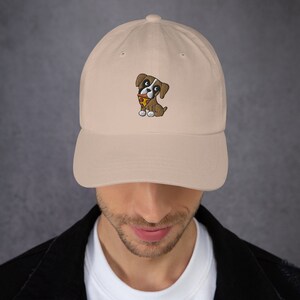 May include: A beige baseball cap featuring an embroidered cartoon dog with a pizza slice. The dog is brown and white with a red bandana. The cap has a curved brim and an adjustable strap.