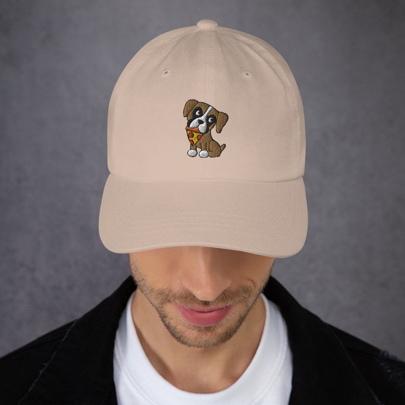 May include: A beige baseball cap featuring an embroidered cartoon dog with a pizza slice. The dog is brown and white with a red bandana. The cap has a curved brim and an adjustable strap.