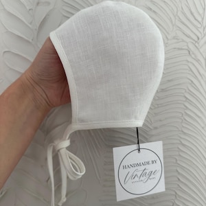 May include: A white linen baby bonnet with ties. The bonnet is held up by a hand. A tag with the words "HANDMADE BY Vintage STYLISH BABY" is attached to the bonnet.