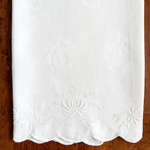 May include: A white rectangular textile featuring a subtle tonal damask floral pattern throughout the fabric. The bottom edge is finished with a scalloped border and detailed white thread embroidery depicting a central floral motif.