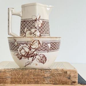 May include: A cream-colored ceramic pitcher and bowl set featuring a brown transferware pattern of roses and geometric lattice designs. The pitcher has an angular handle and a spout, while the bowl has a rounded shape with matching floral motifs.