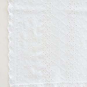 May include: A white fabric textile featuring intricate eyelet embroidery with a repeating floral pattern and vertical bands of stitching. The edges of the material are finished with a scalloped border, creating a delicate and detailed appearance.