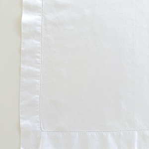 May include: Close-up of a white linen textile with a detailed hem. The fabric has a subtle texture and is bordered by a folded edge with delicate stitching. The overall appearance is clean and crisp, suggesting a high-quality product.