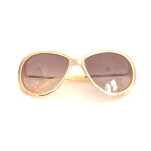 May include: Oversized sunglasses with a gold-toned metal frame and gradient brown lenses. The temples feature a light-colored, patterned design. The brand name Tommy Bahama is printed on the inner side of the left temple arm.