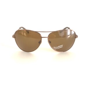 May include: Aviator sunglasses with a metallic bronze frame and brown polarized lenses. The right lens features a small white sticker with the text Tommy Bahama POLARIZED. The design includes a double bridge and thin metal temples.