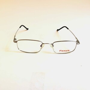 May include: Silver-framed eyeglasses with rectangular lenses and black temple tips. The glasses have a thin metal frame and a nose bridge. The text "Flexure by Capri" is printed in red on the right lens.