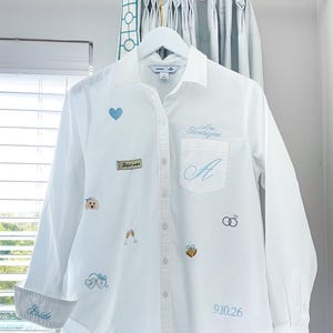 May include: A white button-down shirt featuring light blue embroidery including a heart, a dog, champagne flutes, wedding rings, bells, and the text "Mrs. Fantagini", "A", "Bride", and "9.10.26". A small rectangular patch reads "the perfect match".