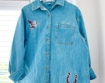 May include: A light blue denim button-down shirt featuring a long-sleeve design, a collar, and a front pocket. The shirt is decorated with an embroidered mallard duck on the right chest, a horseshoe on the lower left, and the initials R.A. above the pocket.