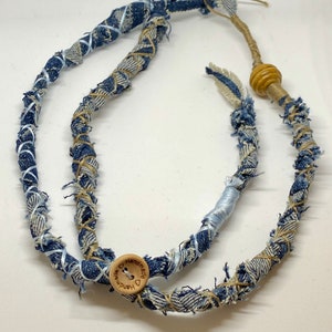 May include: Handmade denim necklace with a unique, rustic design. The necklace features twisted denim fabric in shades of blue and beige, accented with light blue ribbon and a wooden button closure. A wooden bead adds a natural touch.