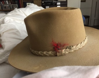 May include: A brown felt cowboy hat with a braided leather band and a small red feather accent.
