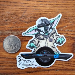 May include: A sticker featuring a cartoon character resembling Baby Yoda wearing a Mandalorian helmet and riding a one-wheeled electric vehicle. The character is wearing a brown coat and has green skin. The vehicle is black with a blue and white logo that says "GT Series".